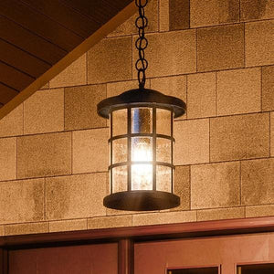 The Urban Ambiance UQL1048 Craftsman Outdoor Pendant Light (15.5"H x 10"W), with a natural black finish and marine-grade construction, hangs by a chain in front of a shingled wall and above a door.