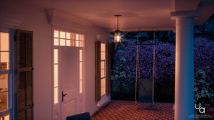 At dusk, a warmly lit porch showcases a white door, shuttered windows, tiled floor, and a ceiling swing with flowering bushes just outside. The space is softly illuminated by the Urban Ambiance UQL1049 Craftsman Outdoor Pendant Light from the Vienna Collection—measuring 15.5″ high by 10″ wide in a Parisian Bronze Finish and crafted with marine-grade construction—adding to the inviting charm of the setting.