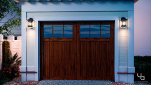 A wooden double garage door with six small seeded glass panes is framed by white siding, brick accents, and two Urban Ambiance UQL1100 Rustic Outdoor Wall Lights in a Black Silk finish for a welcoming touch.