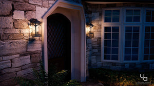 At dusk, a stone house exterior features two Urban Ambiance UQL1102 Rustic Outdoor Wall Lights from the Sydney Collection in black silk finish with seeded glass flanking a dark wooden door, plus large windows on the right.