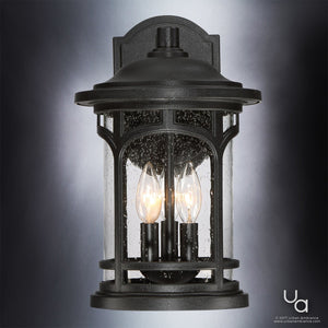 The Urban Ambiance UQL1102 Rustic Outdoor Wall Light from the Sydney Collection features a black silk finish, marine-grade construction, and three candle-style bulbs inside a cylindrical, seeded glass enclosure.
