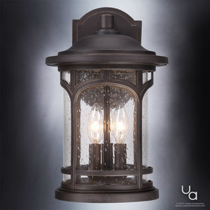 The Urban Ambiance UQL1103 Rustic Outdoor Wall Light from the Sydney Collection features a Parisian Bronze finish, clear seeded glass panels, and two candle-style bulbs. This 14.5-inch high by 9-inch wide marine-grade fixture is shown mounted on a neutral background.