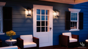 A white door with glass panels is centered on a blue wooden exterior wall, flanked by Urban Ambiance Sydney Collection UQL1104 Rustic Outdoor Wall Lights in black silk finish. Wicker chairs with white cushions and a small table with yellow flowers complete the scene.