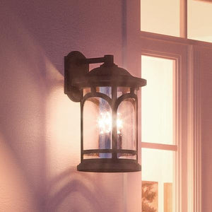The Urban Ambiance UQL1105 Rustic Outdoor Wall Light from the Sydney Collection, featuring a Parisian Bronze finish and marine-grade construction, is mounted next to a window, illuminating the exterior wall at dusk.