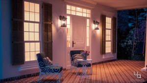 A warmly lit porch at night features two ornate white chairs, a small table with flowers, and a white door flanked by large windows with shutters. Mounted on the wall, the Urban Ambiance UQL1105 Rustic Outdoor Wall Light from the Sydney Collection in a Parisian Bronze finish casts a gentle glow across the scene. Measuring 17.75″ high by 11″ wide and crafted with marine-grade construction, its seeded glass softly diffuses the light for an inviting ambiance.