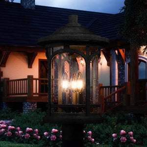 The UQL1106 Rustic Outdoor Post Light by Urban Ambiance, from the Sydney Collection, features marine-grade construction and seeded glass, illuminating a garden at dusk in front of a home with a wooden porch.