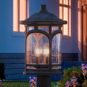The Urban Ambiance UQL1107 Rustic Outdoor Post Light, 19"H x 11"W in Parisian Bronze from the Sydney Collection, featuring marine-grade construction, illuminates a garden near a house at dusk among flowers and greenery.