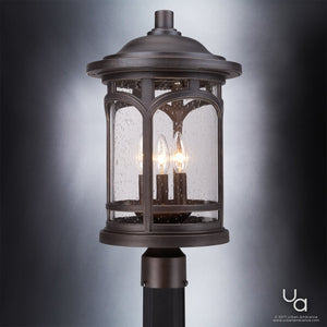 The Urban Ambiance UQL1107 Rustic Outdoor Post Light from the Sydney Collection features a marine-grade construction and is finished in Parisian Bronze. This 19-inch high by 11-inch wide fixture showcases clear seeded glass panels with a raindrop texture and two exposed light bulbs inside, mounted on a post against a gradient gray background.