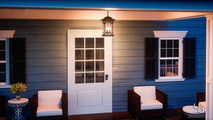 A white door with seeded glass panels is centered between two windows with black shutters, flanked by wicker chairs and a small table topped with yellow flowers on the porch, which is illuminated by the Urban Ambiance UQL1109 Rustic Outdoor Pendant Light from the Sydney Collection in a Parisian Bronze finish.