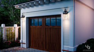 A white garage with a wooden double door features two Urban Ambiance UQL1140 Colonial Outdoor Wall Lights from the Cambridge Collection, each measuring 11.5 inches high by 7 inches wide with a Black Silk finish. The garage is accented by a gravel driveway, sits next to a white fence, and is surrounded by greenery.