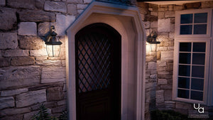 A stone house exterior featuring a dark wooden door, an iron lattice window, and two Urban Ambiance UQL1142 Colonial Outdoor Wall Lights from the Cambridge Collection (14″H x 8″W, Black Silk Finish) illuminated at night, with a white-framed window positioned to the right.