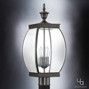 The Urban Ambiance UQL1174 Colonial Outdoor Post Light from the Manchester Collection features a solid brass construction in a Medieval Bronze finish, vintage-transitional styling, and three candle-style bulbs enclosed in clear glass, shown against a gradient gray background.