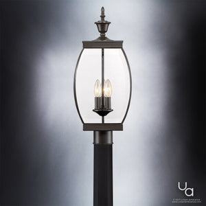 The Urban Ambiance UQL1174 Colonial Outdoor Post Light from the Manchester Collection features a medieval bronze finish and stands 22 inches high by 9 inches wide. This vintage-transitional fixture showcases a clear glass lantern enclosure with solid brass construction and three candle-style bulbs, presented elegantly against a neutral gradient background.