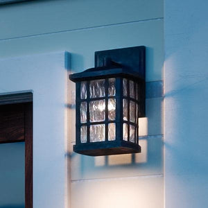 The Urban Ambiance UQL1230 Zurich Collection Craftsman Outdoor Wall Light, featuring a black silk finish and marine-grade construction, is mounted on a light exterior wall and emits a soft glow at dusk.