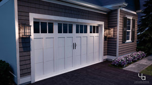 A white garage door featuring windows at the top is flanked by Urban Ambiance UQL1235 Craftsman Outdoor Wall Lights from the Zurich Collection, each measuring 15.5 inches high by 9.5 inches wide and finished in Parisian Bronze with marine-grade construction for durable, weather-resistant outdoor lighting, alongside a flower bed bordering the house.