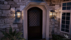 A black UQL1236 Craftsman Outdoor Wall Light by Urban Ambiance illuminates a stone wall entry with a diamond glass door, its marine-grade Zurich design flanked by a window and leafy plant for striking architectural accent.