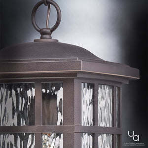 Close-up of the Urban Ambiance UQL1251 Craftsman Outdoor Pendant Light from the Zurich Collection, measuring 15 inches high by 9.5 inches wide, in a Parisian Bronze finish with marine-grade construction. The fixture features textured water glass diffuser panels and a metal ring at the top, set against a gradient gray background.