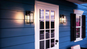 A white glass-paneled door with a black handle on a blue house is flanked by two Urban Ambiance UQL1330 Farmhouse Outdoor Wall Lights from the Quincy Collection, featuring a black finish and beveled glass, next to a window with black shutters at sunset.