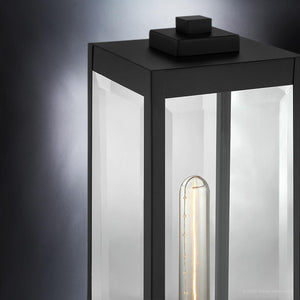 Close-up of the Urban Ambiance UQL1336 Modern Farmhouse Outdoor Pier Light from the Quincy Collection, showcasing a 16-inch tall, 7.5-inch wide black steel fixture with clear beveled glass panels and a visible vertical bulb inside, set against a gradient gray background.