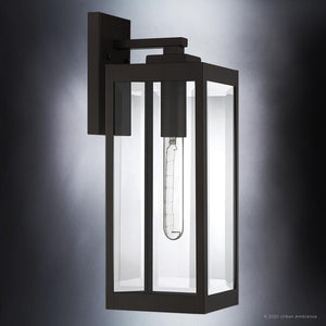 The Urban Ambiance UQL1351 Modern Farmhouse Outdoor Wall Light from the Quincy Collection features a rectangular black metal frame with clear glass panels and a visible tubular bulb, all mounted on a square wall plate against a gray gradient background. Measuring 17 inches high by 6 inches wide, this striking outdoor wall sconce seamlessly combines function with modern farmhouse style, while its Estate Bronze finish lends a refined, contemporary touch to any exterior.