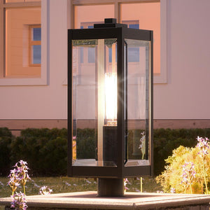 At dusk, the Urban Ambiance UQL1353 Modern Farmhouse Outdoor Post/Pier Light from the Quincy Collection, featuring an Estate Bronze finish and clear beveled glass panels, is illuminated atop a stone base in a garden with purple flowers before a house with glowing windows—showcasing the perfect blend of modern farmhouse outdoor lighting.