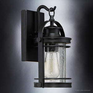 The Urban Ambiance UQL1430 Nautical Outdoor Wall Light from the Cannes Collection features a Black Silk finish, an 11.5″ high by 6.75″ wide metal frame with a cylindrical seedy glass shade and visible bulb, mounted on a rectangular backplate—a vintage-inspired design that seamlessly combines classic charm and modern durability.
