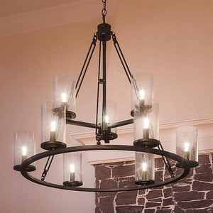 A UQL2131 Industrial Chandelier from the Valencia Collection by Urban Ambiance, featuring an estate bronze finish and eight glass-shaded candle-style bulbs, hangs from the ceiling in a room with stone and beige walls.