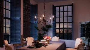 A dining room with wood-tone accents features the Urban Ambiance UQL2350 Americana Chandelier from the Raleigh Collection, showcasing a rustic bronze finish and measuring 14.25″H x 24″W, hanging above a table adorned with dishes and flowers. The space is illuminated by evening light streaming through large windows framed by dark shutters.