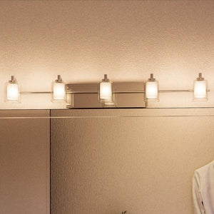 A bathroom wall features a large mirror with four Urban Ambiance UQL2407 Modern Bathroom Vanity Lights from the Napa Collection, each in a polished chrome finish with frosted glass shades, installed above it; on the right, there is a partial view of a person in a white shirt.