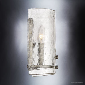 The Urban Ambiance UQL2873 Industrial Wall Sconce from the Nantes Collection features a textured glass shade and a visible bulb, casting a soft glow against a gray background. This wall-mounted fixture measures 10″ high by 8.75″ wide and is finished in Silver Etch.