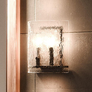The Urban Ambiance UQL2873 Industrial Wall Sconce from the Nantes Collection, featuring a curved, textured glass shade and silver etch finish metal fixtures, is mounted on a tiled wall beside a wooden frame.