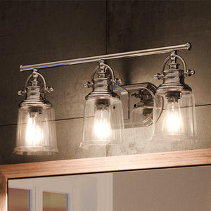 The Urban Ambiance UQL2881 Industrial Bathroom Vanity Light from the Salford Collection features three clear glass shades and a polished chrome finish, mounted above a mirror on a concrete wall to provide sleek industrial bath lighting with a modern flair. The fixture measures 9.5 inches high by 23 inches wide.