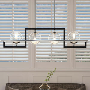 Urban Ambiance UQL3241 Minimalist Chandelier from the Odessa Collection, featuring a natural black rectangular frame with four glass globe bulbs, is suspended in front of white window shutters above a gray couch with a glass vase.