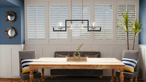 A dining area featuring a wooden table, two gray chairs with striped pillows, a bench, a lantern centerpiece, potted plant, round mirrors, and expansive white shuttered windows is elegantly illuminated by the Urban Ambiance UQL3241 Minimalist Chandelier (10.5″H x 40″W) in Natural Black Finish from the Odessa Collection, adding a stylish and contemporary touch to the space.