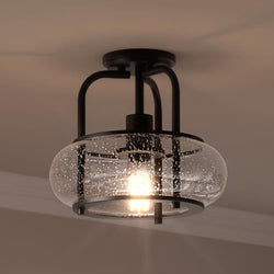 The UQL3281 Utilitarian Ceiling Light from Urban Ambiance's Clearwater Collection is mounted on a beige ceiling. It features a dark bronze frame with a spherical glass cover that has a bubbled texture reminiscent of vintage-inspired bulbs. Dimensions: 10"H x 12"W.