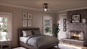 A cozy bedroom includes a brick fireplace with vintage bulbs, bed with gray bedding, and a potted plant. Fern artwork and photos decorate the mantel. Natural light flows through two windows beneath the Urban Ambiance UQL3281 Utilitarian Ceiling Light in Dark Bronze from the Clearwater Collection.
