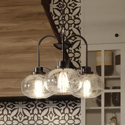 The UQL3351 Utilitarian Chandelier from Urban Ambiance's Clearwater Collection features vintage-inspired bulbs within oversized clear seedy glass shades, perfect for illuminating rooms with decorative black and white patterned tiles and wooden cabinetry. It has a 16.5"H x 22"W dark bronze finish.