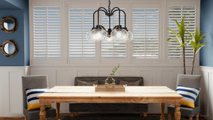A dining room showcases a wooden table, gray chairs, and a bench by white-paneled windows. The Urban Ambiance UQL3361 Utilitarian Chandelier with vintage-inspired bulbs hangs above. Blue walls feature round mirrors, and a tall plant adds charm to the corner.