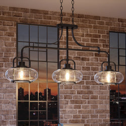 The Urban Ambiance UQL3381 Utilitarian Chandelier, part of the Clearwater Collection with a dark bronze finish measuring 21.55"H x 38"W, features vintage-inspired bulbs and three oversized clear seedy glass shades, offering a captivating view against a brick wall.