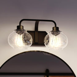 The UQL3402 Utilitarian Bath Vanity Light from Urban Ambiance's Clearwater Collection features two decorative bubble glass shades above a curved mirror and vintage-style bulbs, all in a Dark Bronze finish.