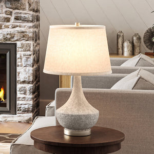 A gorgeous UEX7360 New Traditional Table Lamp in a polished concrete finish, providing luxurious lighting on a table next to a fireplace.