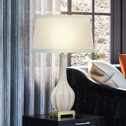 A beautiful UEX7390 Traditional Table Lamp by Urban Ambiance, with an Aged Brass and White Finish, providing luxury lighting on a table next to a window.