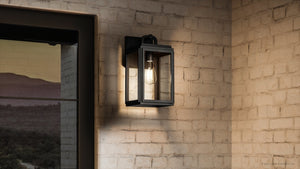 The Urban Ambiance UHP1381 Farmhouse Outdoor Wall Sconce from the Macon Collection features a marine-grade, corrosion-resistant olde bronze finish and clear glass, ensuring lasting beauty outdoors; shown wall-mounted at dusk beside a window.