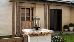 The UHP1384 Farmhouse Outdoor Post Light by Urban Ambiance, 15.625''H x 7''W in Olde Bronze with marine-grade construction, stands on a white brick pillar in front of double glass doors and a garden with flowers and potted plants.