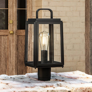 The UHP1384 Farmhouse Outdoor Post Light by Urban Ambiance, featuring an Olde Bronze finish and marine-grade construction, stands on a stone surface before a wooden door and brick wall.