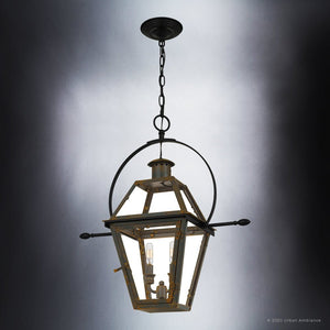 The UQL1378 Historic Outdoor Pendant Light from Urban Ambiance’s Paris Collection features a Bygone Bronze finish, black metal frame with glass panels, and three visible light bulbs, evoking the look of a handcrafted colonial gas lantern. Measuring 23.5″ high by 21.25″ wide, this fixture is suspended by a chain from a ceiling mount against a neutral background.