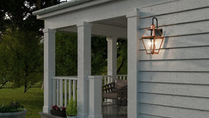 The Urban Ambiance UQL1382 Historic Outdoor Wall Light from the Paris Collection, featuring a rustic copper finish and measuring 22.5″ high by 10.5″ wide, emits a warm glow on the exterior wall of a light gray porch accented by white columns, railing, and a few plants in the foreground.