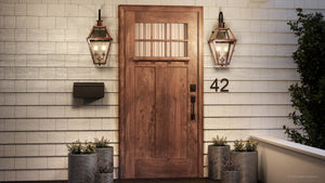 A welcoming entryway is created with a wooden front door featuring glass panels, flanked by two Urban Ambiance Paris Collection UQL1382 Historic Outdoor Wall Lights in a rustic copper finish (22.5″H x 10.5″W), complemented by a handcrafted copper lantern, mailbox, the number 42, and potted plants against a white shingle wall—blending vintage outdoor lighting charm with contemporary elegance.