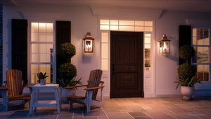 A warmly lit front porch with two wooden chairs, a small table, potted plants, a large wooden door, and two handcrafted Urban Ambiance UQL1411 Vintage Outdoor Wall Lights (23″H x 12″W, Rustic Copper Finish, Longview Collection) in the style of vintage lantern lights.