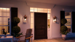 At dusk, a front porch features a wooden door, two potted plants, a wooden chair, large windows, and the Urban Ambiance UQL1413 Vintage Outdoor Wall Light from the Longview Collection in a rustic copper finish.